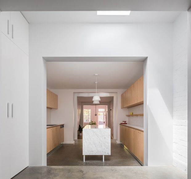 Bright, minimalist galley kitchen viewed through a wide opening. White full-height cabinetry and walls frame warm timber lower and upper cabinets, with a central marble-clad island, dark polished concrete flooring, and a simple pendant light. A dining or living area is visible beyond the kitchen, while sunlight falls across the white brick wall at right.