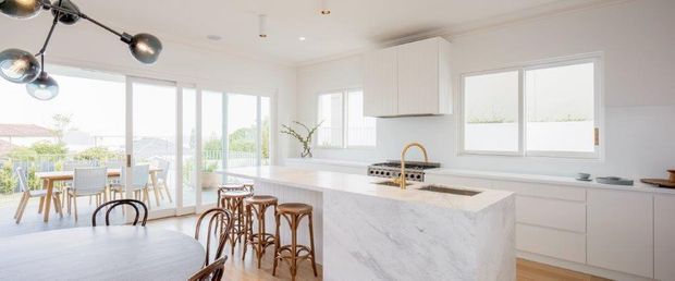 Bright, open-plan contemporary kitchen and dining area with a large white marble-look island, integrated white cabinetry, timber bar stools, and a dining table beside full-height glazed doors opening to an outdoor area. The space has abundant natural light and a restrained neutral palette, with a sculptural black pendant light as a focal point.