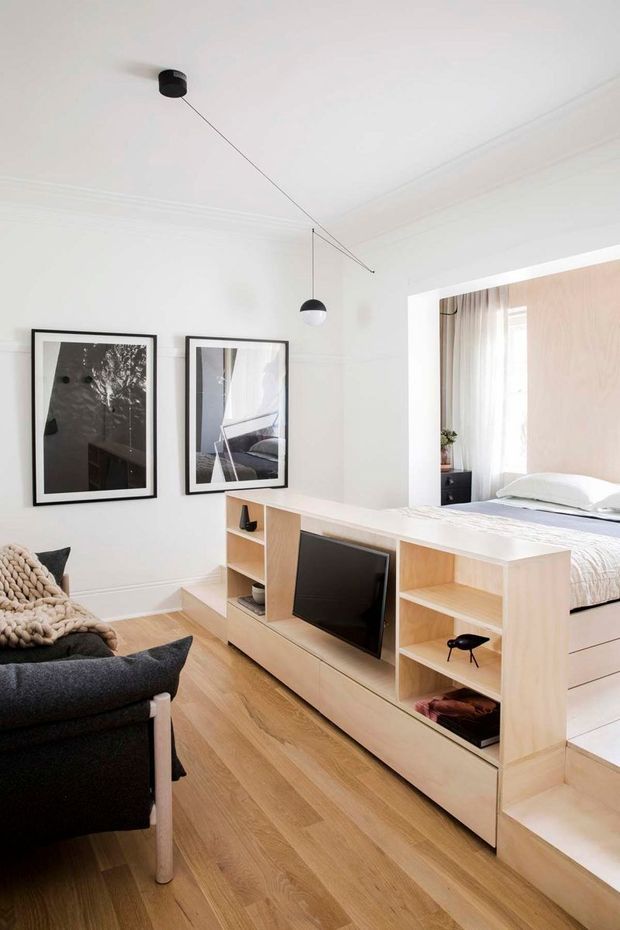 Bright, compact open-plan living space with a built-in pale timber media unit and room divider separating the living area from a bedroom. The room features white walls with decorative cornice detailing, light timber flooring, framed monochrome artwork, a dark upholstered armchair, and minimalist suspended pendant lighting. The bedroom beyond has pale textured wall panelling, white bedding, and soft curtains.