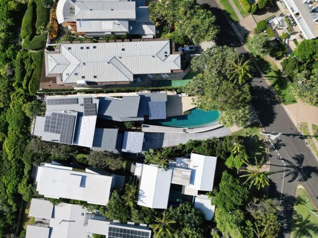 Aerial top-down view of a leafy suburban residential neighbourhood, showing several detached homes with predominantly light-coloured metal roofs, dense gardens, solar panels, and a long narrow turquoise swimming pool beside one of the central properties. A paved road and footpaths run along the right side of the image.