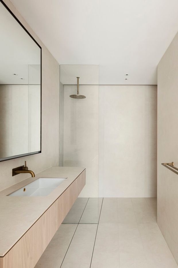 A bright, minimalist bathroom with a long floating timber-veneer vanity, integrated pale stone benchtop and white basin. A large black-framed mirror spans the left wall, while a frameless glass shower enclosure with a round rainfall showerhead occupies the rear. Large pale floor and wall tiles create a calm, cohesive interior, with brushed brass tapware and hardware providing subtle accents.