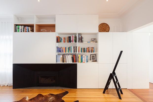 Bright, refined living room with a predominantly white built-in media and storage wall incorporating open bookshelves and concealed cabinetry. A dark fireplace surround creates a strong focal point against polished timber flooring, with a minimalist easel-like floor object and restrained decorative accents. Traditional ceiling cornices and the timber floor contrast with the contemporary custom joinery.