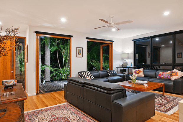 A spacious, bright living room with black leather sectional seating arranged around a wooden coffee table, layered patterned rugs, and timber flooring. Large timber-framed glazed doors and windows open the room toward lush tropical greenery and a deck. The room includes a ceiling fan, recessed lighting, dark window treatments, and warm wood accents.