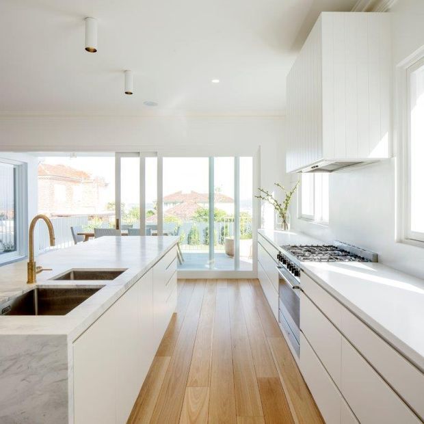 Bright contemporary white kitchen with a long central island, integrated double sink and brass tap, white cabinetry, a white stone island benchtop, stainless-steel cooking appliances, and warm timber flooring. Full-height glazing at the end of the room opens to a balcony or exterior view, creating an airy feel.