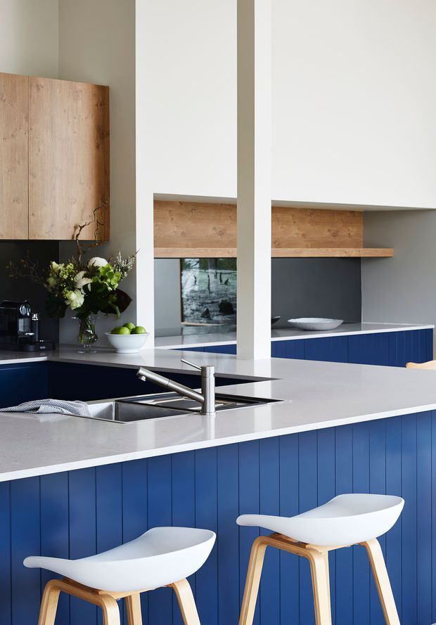 Bright contemporary kitchen with a large white stone island, deep blue vertical-panel cabinetry, pale timber overhead cabinetry and shelving, dark splashback areas, and two white-and-timber bar stools. A sink and tap are integrated into the island, with a coffee machine, fruit bowl, and floral arrangement on the rear bench.