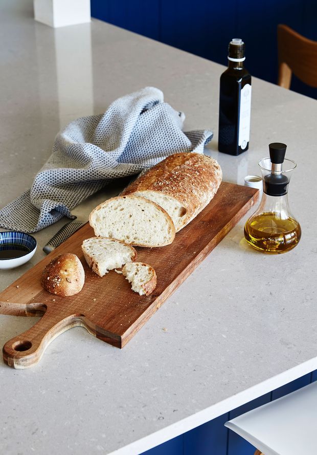 Close-up of a styled kitchen island with a light grey stone benchtop, featuring a wooden cutting board with sliced artisan bread, a linen cloth, olive oil bottles, and a small bowl. Deep blue cabinetry or paneling and a timber chair are visible in the background.