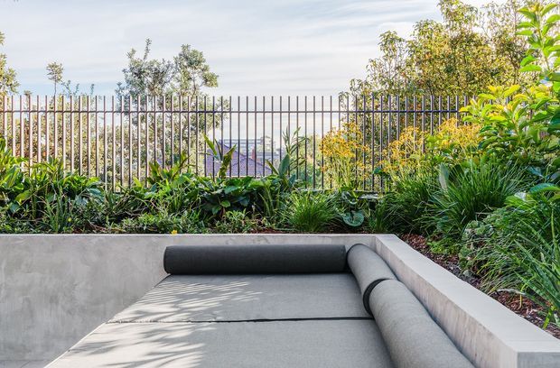 A landscaped outdoor lounge with a built-in L-shaped concrete seating area and grey upholstered cushions, surrounded by dense planting. A slim vertical metal fence lines the rear boundary, with a distant urban or coastal view visible beyond it. The scene has a contemporary, relaxed garden atmosphere with strong natural greenery and filtered sunlight.