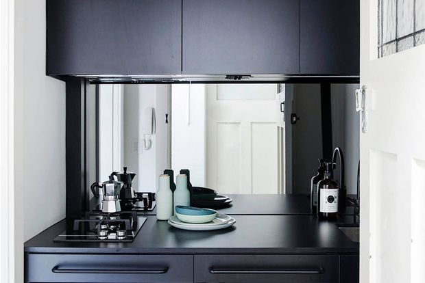 Compact, dark-toned kitchen or kitchenette with a black built-in benchtop and cabinetry, integrated gas cooktop, and mirrored splashback reflecting a white interior door. The worktop is styled with a stovetop espresso maker, ceramic vessels, stacked dishes, and a soap dispenser. White walls and a small high window provide contrast and natural light.