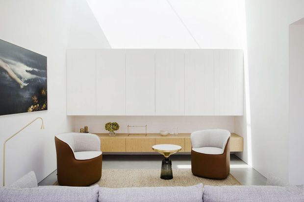 Bright, minimalist living room with a low built-in timber media or storage console, extensive white overhead cabinetry, two sculptural brown-and-white lounge chairs, a small round coffee table, and a pale sectional sofa in the foreground. The room has a high sloping white ceiling, polished grey floor, neutral textured rug, contemporary artwork, and a slim floor lamp.