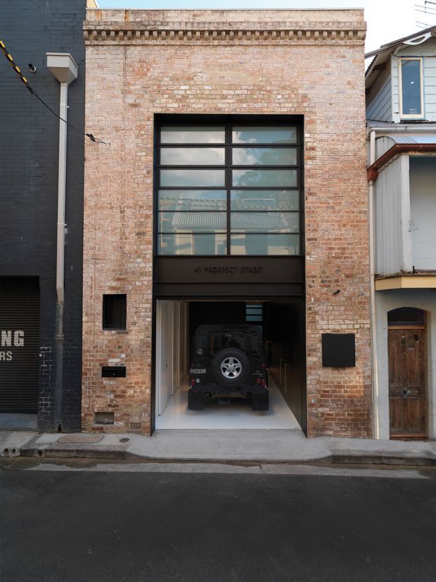 Street-facing narrow brick building facade with a weathered pale-brick exterior, decorative cornice detailing, a large black-framed multi-pane window, and a wide ground-level garage opening containing a vehicle. The building is tightly flanked by neighbouring urban structures, with a small street frontage and visible overhead utility lines.
