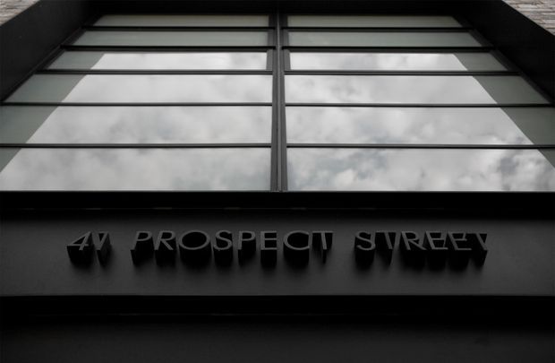 Close-up exterior view of a dark commercial building facade, featuring reflective horizontal window panels and raised black lettering reading “4V Prospect Street.” The image has a monochromatic, high-contrast architectural photography style.