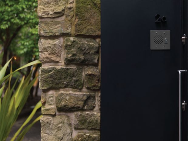 Close-up exterior view of a dark matte entry door beside a rustic, weathered stone-and-mortar wall. A small metal intercom or access panel and door hardware are visible, with lush planting along the adjacent walkway.