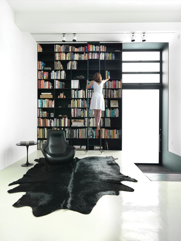 Bright contemporary reading room dominated by a floor-to-ceiling black built-in bookcase filled with books. A person in a white dress reaches toward the shelves using a small ladder. The room has white walls and ceiling, a large black-framed glazed door or window, polished light flooring, a black leather lounge chair, a small round side table, and a dark irregular hide rug. Track lighting runs along the ceiling.
