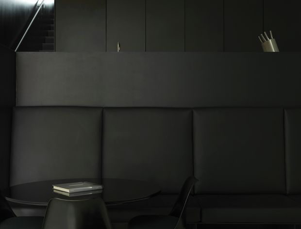 A dark, minimalist interior showing a built-in black upholstered banquette or sectional seating against a charcoal wall. A small dark table with stacked books sits in the foreground, with black vertical panelled cabinetry or wall cladding behind the seating and a partially visible angular staircase or architectural element at upper left. The composition is moody, monochromatic, and refined.
