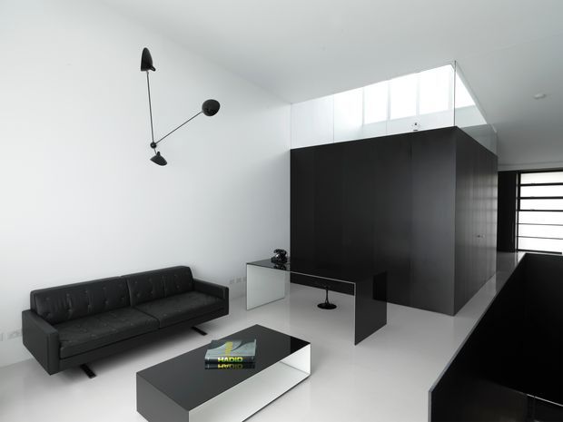 Minimalist open-plan living room with a black leather sofa, low black-and-white coffee table, sculptural wall light, and a large dark built-in joinery volume. White walls and flooring, high clerestory glazing, and a black-framed horizontal window create a stark monochrome interior.