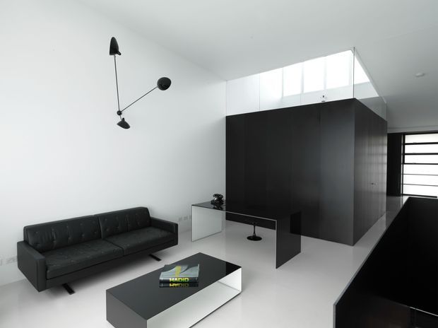 Minimalist open-plan interior with a black leather sofa, low black-and-white coffee table, sculptural wall light, dark built-in cabinetry or room divider, and a partially visible kitchen counter. White walls, pale flooring, and a high clerestory window create a stark monochromatic composition.