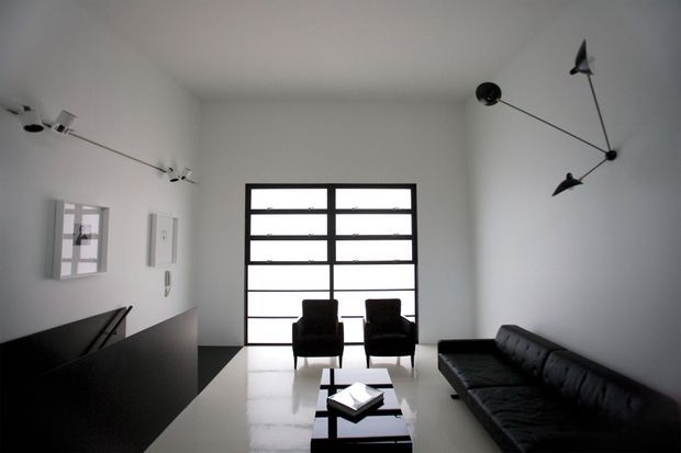 Minimalist living room with a black leather sofa, two black lounge chairs, a low black coffee table, and a dark built-in cabinet or partition. White walls and a glossy pale floor create a stark monochrome scheme. A large black-framed glazed door or window fills the rear wall, while sculptural directional wall and ceiling lighting adds a distinctive mid-century-inspired detail.