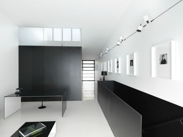 A bright, minimalist black-and-white interior resembling a gallery or creative studio. The long narrow space features a white polished floor and walls, a high white ceiling with track lighting, black built-in cabinetry or partitions, a compact desk with an office chair, framed monochrome artworks, and a glazed rear door or partition allowing daylight into the space.
