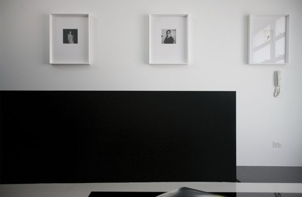 Minimalist interior view dominated by a long matte-black partition or media wall, with a reflective dark surface in the foreground. White walls feature three simple white-framed artworks and a wall-mounted intercom. The restrained monochrome composition has clean lines and sparse decoration.