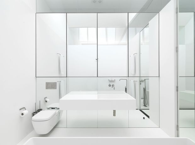 Bright, minimalist white bathroom viewed from the bathtub area, featuring a wall-hung toilet, floating double vanity with integrated basin, mirrored and white cabinetry, chrome fixtures, and a glass-enclosed shower. The room has clean linear detailing, abundant natural light, and a highly monochromatic finish.