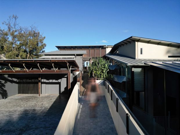 Exterior architectural view of a contemporary multi-level residential complex, showing a covered carport at left and a narrow elevated pedestrian walkway leading between connected buildings. The buildings feature low-pitched metal roofs, dark metal framing, rendered light walls, timber-toned cladding, and extensive glazing. Clear blue sky and established trees surround the structure.