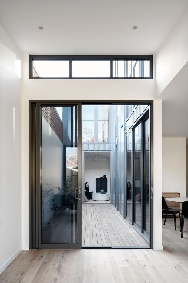 A bright contemporary interior looking through a large dark-framed glazed opening onto a narrow internal courtyard or lightwell. Pale timber flooring, white walls and ceiling, clerestory windows, and extensive black-framed glazing create a clean, airy connection between the interior and courtyard. A dining area with a table and dark chairs is partially visible on the right, while the courtyard beyond contains pale decking, vertical metal cladding, brickwork, and a small seating area.