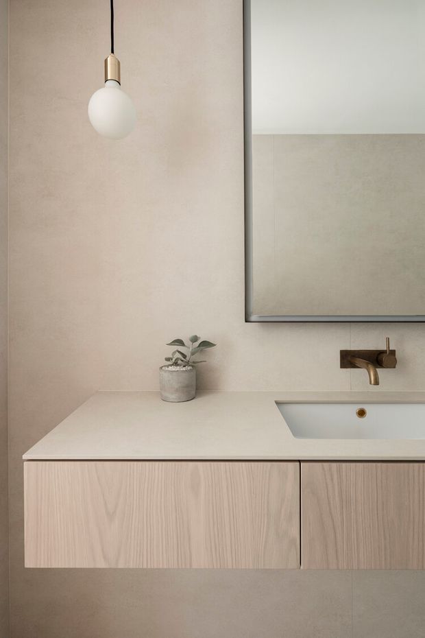 A minimalist bathroom vanity with a pale engineered stone benchtop, integrated white basin, light timber floating cabinetry, and a wall-mounted aged brass tap. The warm beige plastered walls, large simple mirror, spherical pendant light, and small potted plant create a calm, refined and softly monochromatic interior.