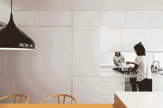 A bright, minimalist contemporary kitchen and dining area with extensive white, handleless cabinetry, a large white island or benchtop in the foreground, integrated cooking appliances, and a reflective backsplash or mirror showing two people preparing food. A glossy black pendant light hangs above the dining table, with timber dining chairs visible at the lower edge.