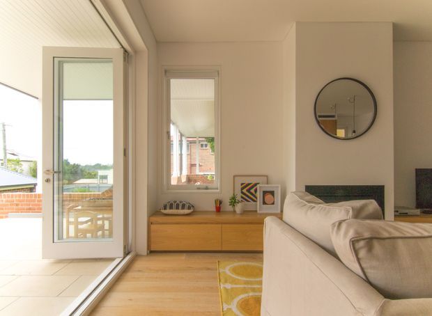 Bright contemporary living room with pale timber flooring, a low timber media console, cream sofa, and a round wall mirror. Large glazed doors open to an outdoor terrace, while a narrow window brings in additional daylight. The room has warm neutral finishes, simple built-in elements, and colourful geometric artwork and rug accents.