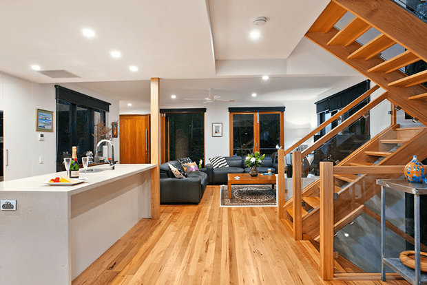 Open-plan residential interior showing a contemporary kitchen with a white island benchtop, timber flooring, living area, dark-framed windows, and a prominent timber staircase with glass balustrades.