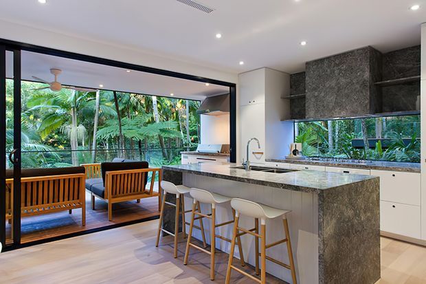 A bright, open-plan contemporary kitchen with a large stone island and four white bar stools, integrated cabinetry, dark textured upper cabinetry, and a wide splashback window. Black-framed sliding glass doors open directly to a furnished outdoor area surrounded by lush tropical vegetation. The space has pale timber flooring, recessed ceiling lighting, and a clean indoor-outdoor connection.