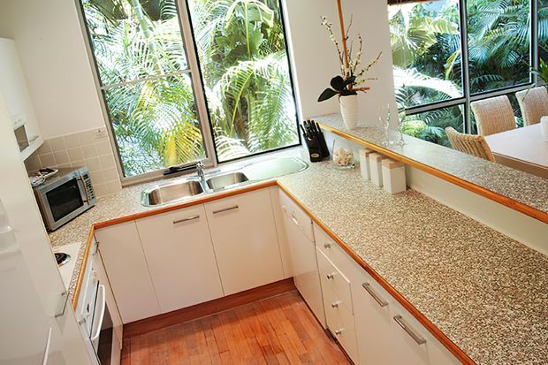 Bright compact U-shaped kitchen with cream fixed cabinetry, integrated appliances, stainless-steel sink, and extensive speckled stone-look benchtops. Large black-framed windows overlook lush tropical foliage, while a raised peninsula with bar seating connects to the adjacent area. Warm timber flooring and orange-toned benchtop edging add contrast.