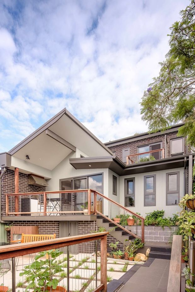 Exterior view of a multi-level contemporary brick-and-cladding home arranged around landscaped terraces and stairs. The facade combines dark brown brick, pale vertical cladding, broad glazed windows, projecting roof forms, and timber-toned metal balustrades. Paved paths, raised planters, potted plants, and outdoor deck areas create a layered courtyard-like setting under a bright partly cloudy sky.