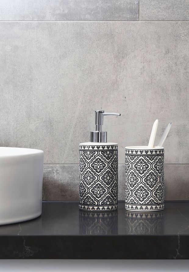 Close-up bathroom vanity scene featuring a white basin, chrome single-lever tap, and matching black-and-white patterned ceramic soap dispenser and toothbrush holder on a dark polished benchtop. Large grey concrete-look wall tiles form the backdrop.