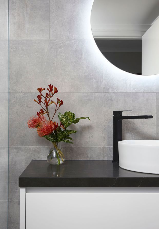 Close-up view of a contemporary bathroom vanity with large light grey tiled walls, a black stone benchtop, white integrated vanity cabinetry, a white vessel basin, and matte black tapware. A round backlit mirror and a small floral arrangement add decorative detail.