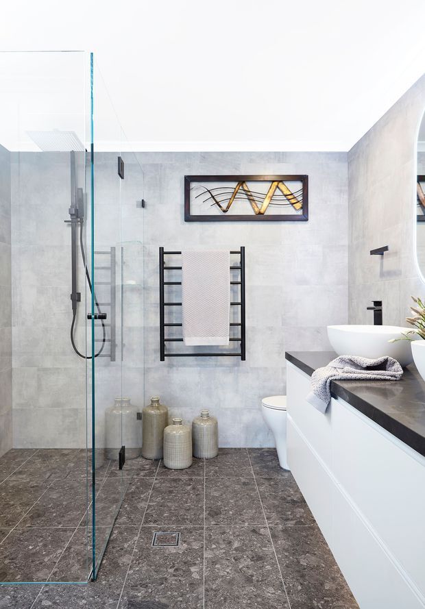 Bright contemporary bathroom featuring a frameless glass walk-in shower, grey stone-look wall and floor tiles, a white freestanding vessel bathtub, and a white floating vanity with a dark stone benchtop. Black tapware, shower fittings, heated towel rail, decorative ceramic vessels, and abstract wall art add contrast.