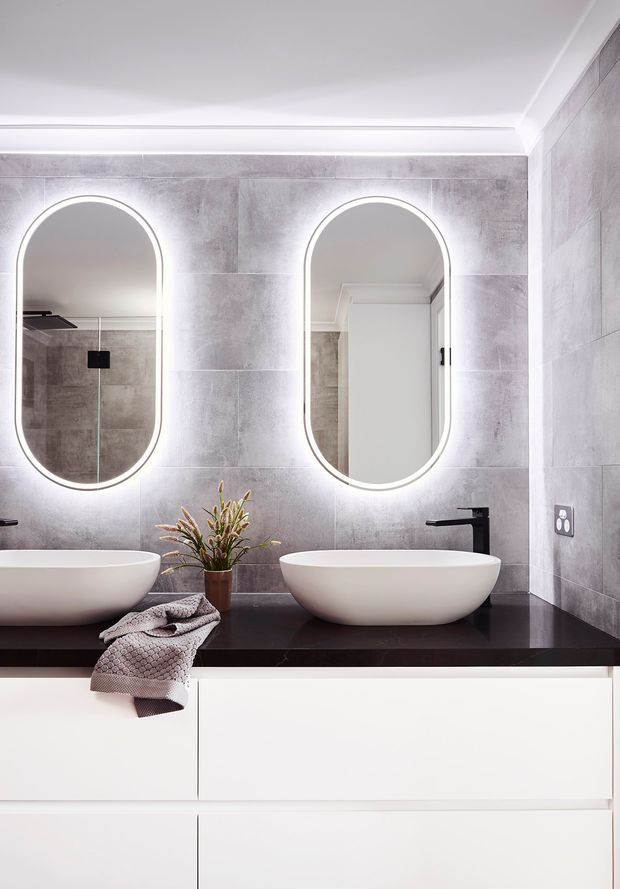 Contemporary bathroom vanity with two white vessel basins on a dark benchtop, white integrated cabinetry, grey large-format wall tiles, black tapware, and illuminated arched mirrors.