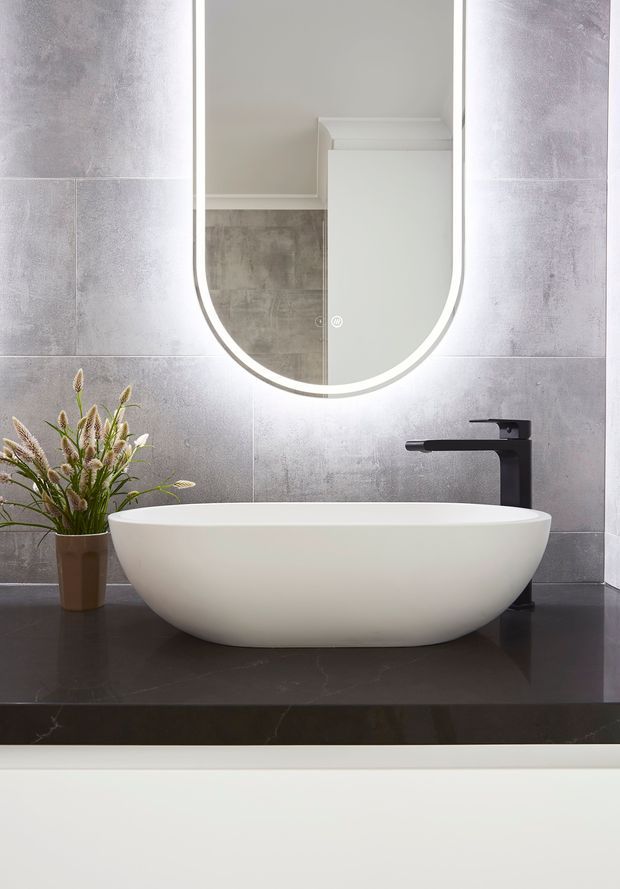 A contemporary bathroom vanity vignette featuring a white freestanding vessel basin set on a dark stone benchtop, with matte black tapware and a rounded illuminated mirror. Large grey concrete-look wall tiles create a textured backdrop, with a small plant in a brown pot adding a natural accent.