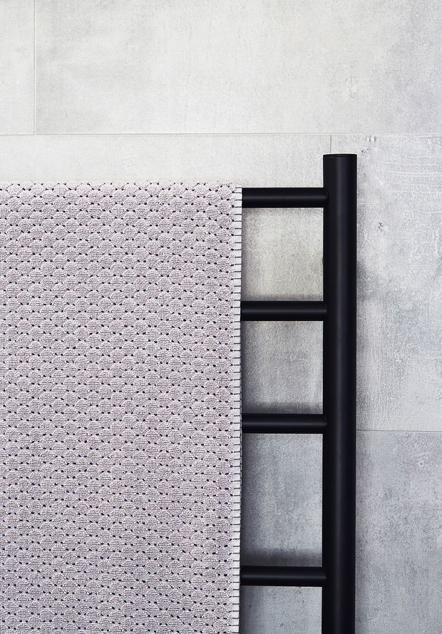Close-up of a contemporary bathroom heated towel rail in matte black, with a pale textured towel draped over the horizontal bars. The rail is mounted against large light-grey concrete-look wall tiles, creating a minimal, monochromatic bathroom detail.