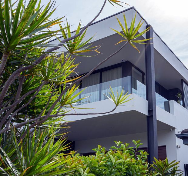 A contemporary multi-storey residential exterior with crisp white and charcoal geometric volumes, deep roof overhangs, projecting balconies, and extensive glazing. Lush tropical planting frames the foreground and partially obscures the lower level.