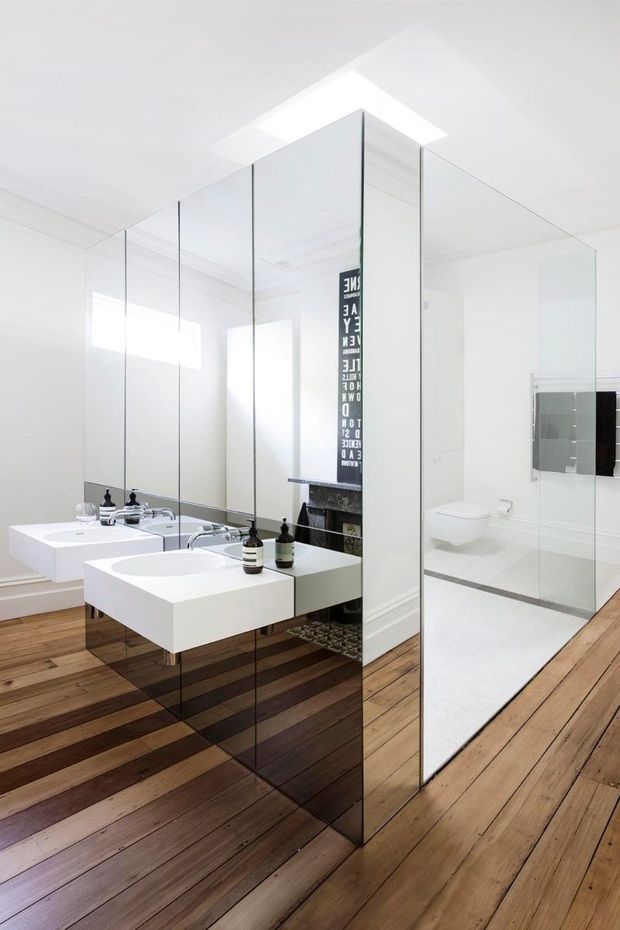 Bright contemporary bathroom with a double floating vanity, wall-mounted basins, chrome tapware, large frameless mirrored panels, and a glass-enclosed toilet area. The room has white walls and sanitaryware, timber plank flooring, and a skylight that provides strong natural illumination. A dark vertical artwork is reflected in the mirrors.