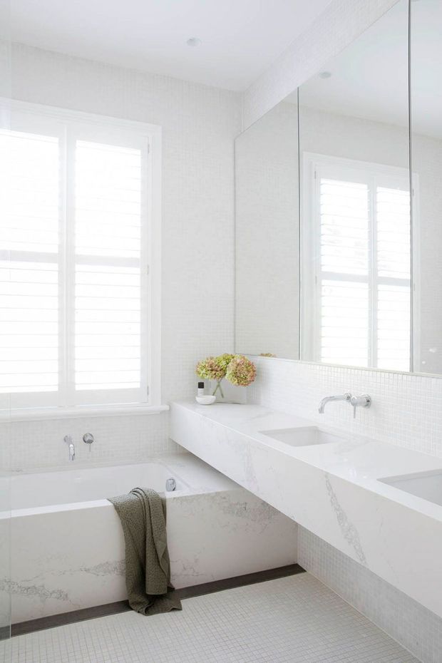 Bright, predominantly white contemporary bathroom with a freestanding bathtub, long double vanity, wall-mounted taps, large frameless mirror, white mosaic wall and floor tiles, and white plantation shutters. Subtle veined stone surfaces and a muted towel add restrained contrast.