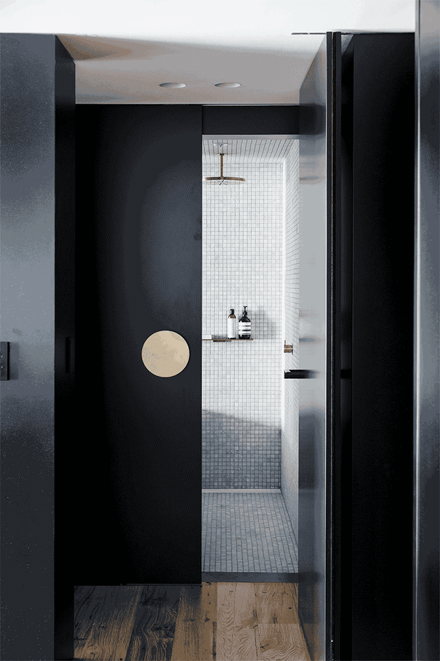 A narrow, moody bathroom viewed through a partially open matte-black door. The shower enclosure is finished in small pale grey mosaic tiles on the walls and floor, with a ceiling-mounted round showerhead, wall-mounted shelf, and dark fixtures. Black built-in door and joinery contrast with warm timber flooring outside the shower.