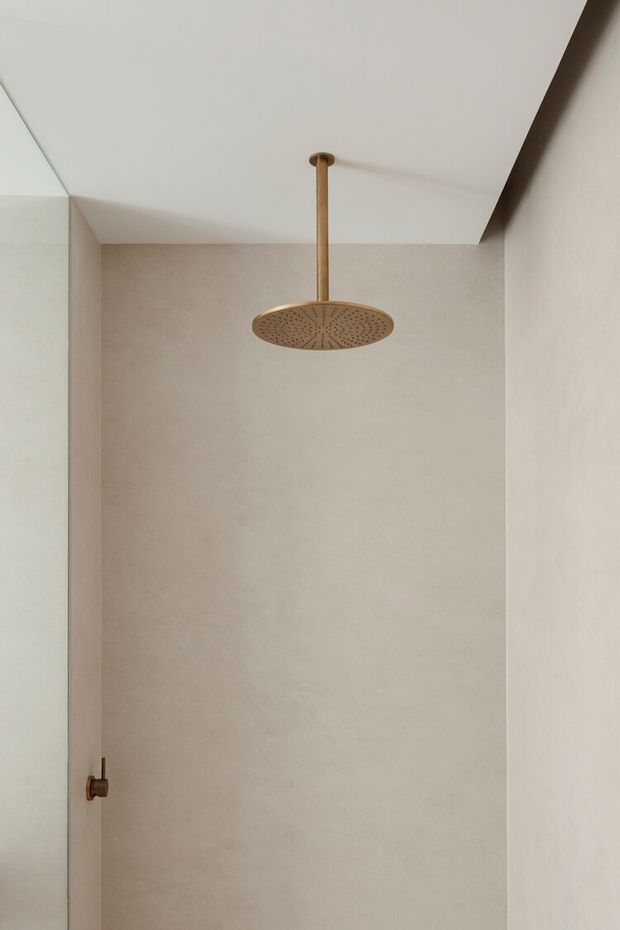Minimal walk-in shower enclosure with large-format warm beige wall tiles, a white ceiling, clear glass partition on the left, and a round brushed brass rainfall showerhead with matching brass controls. The space is uncluttered and softly lit.