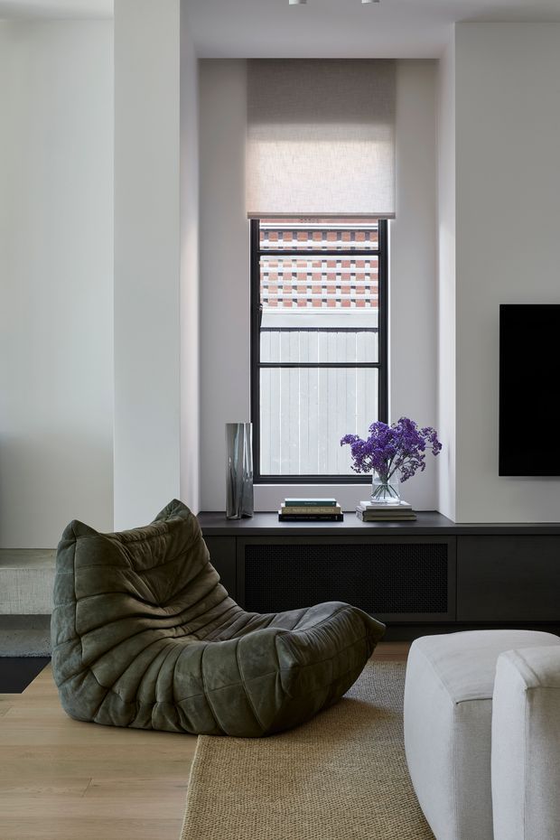 A refined contemporary living room with a tall recessed window, roller blind, dark built-in media cabinetry, and wall-mounted television. A sculptural olive-green lounge chair and white upholstered sofa sit on a natural woven rug over pale timber flooring. Books, a vase of purple flowers, and decorative objects style the low console. The room has a restrained, soft-neutral palette with strong dark joinery and black window framing.