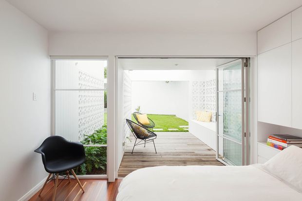 Bright, minimalist bedroom opening directly onto a timber deck and enclosed courtyard garden. The room has white built-in cabinetry, white walls, a low-profile bed, polished timber flooring, a black sculptural chair, and large glazed doors creating a strong indoor-outdoor connection. A small lawn and patterned masonry privacy walls are visible beyond the deck.