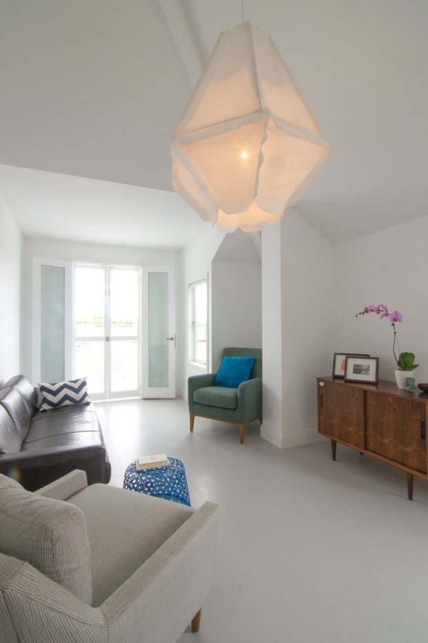 Bright contemporary living room with white walls and flooring, large glazed doors, a grey sofa and armchair, a wooden mid-century-style sideboard, and a sculptural pendant light. Blue cushions and a small patterned ottoman provide colour accents.