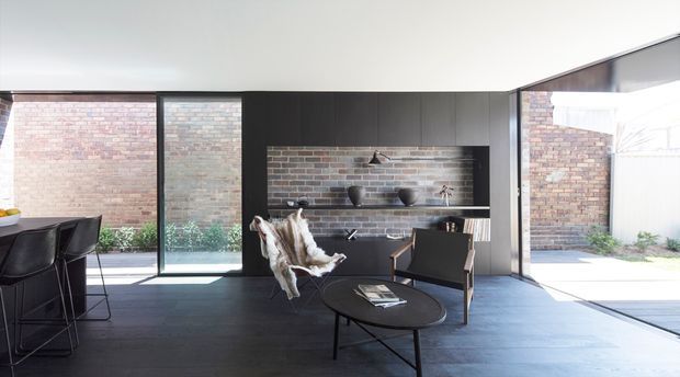 A refined contemporary living and dining area with dark timber-toned built-in cabinetry, a black tiled feature wall and open shelving, dark flooring, and expansive floor-to-ceiling glazing. A compact dining setting is visible at left, while a lounge arrangement with a black round coffee table, armchair, and sculptural pendant occupies the foreground. Exposed brick courtyard walls are visible through the glazing, creating a strong indoor–outdoor connection.