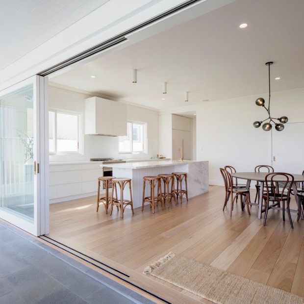 Bright open-plan kitchen and dining area viewed from a covered outdoor threshold through wide sliding glass doors. The room features white cabinetry, a pale stone island with timber bar stools, light timber flooring, a round dining table with bentwood-style chairs, and a sculptural dark pendant light. The space has a clean, airy contemporary coastal character.
