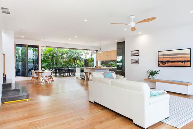 Bright open-plan living, dining and kitchen area with white walls and ceiling, warm timber flooring, large glazed doors opening to lush greenery, a white sectional sofa, timber dining furniture, and a kitchen island with light timber cabinetry. A ceiling fan, recessed lighting, and restrained contemporary furnishings create an airy indoor-outdoor feel.